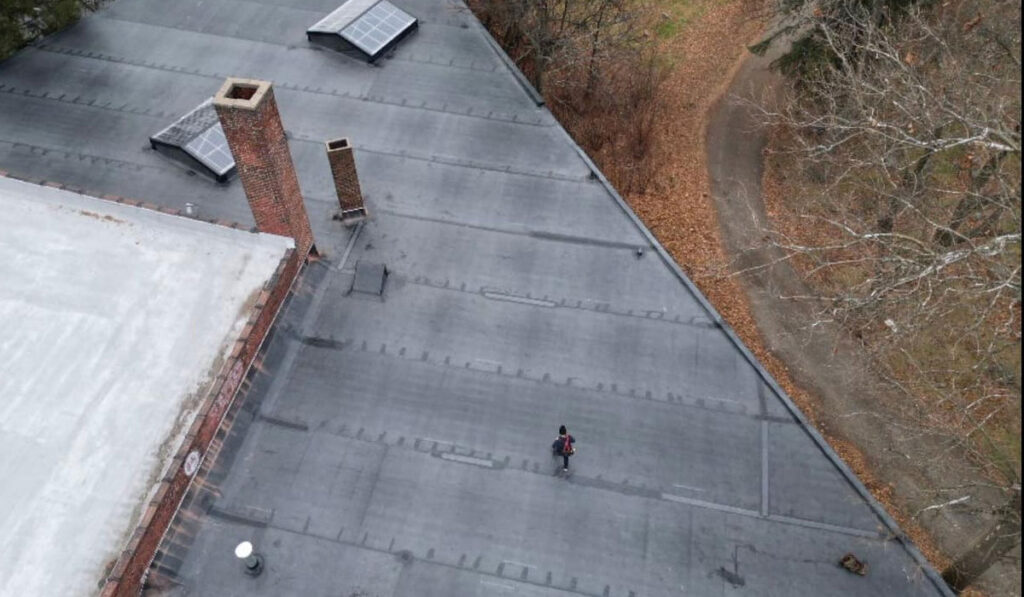 Flat commercial roof with a person inspecting drainage, visible chimneys, and solar panels, highlighting maintenance and ponding prevention.