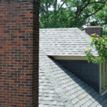 Roofing structure with shingles adjacent to a brick chimney, surrounded by greenery, illustrating residential roofing options and aesthetic appeal.