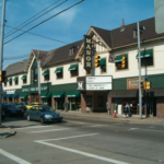Street view of the Manor building in Stanton Heights, Pittsburgh, featuring green awnings, traffic lights, and pedestrians, illustrating the neighborhood's commercial area relevant to local roofing maintenance concerns.