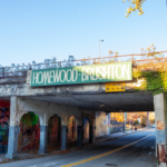 Urban underpass with "Homewood-Brushton" sign, surrounding graffiti, and clear sky, symbolizing community in Pittsburgh's neighborhoods.