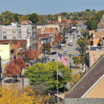 View of a tree-lined street in Carrick, showcasing autumn foliage, local businesses, and an American flag, emphasizing the community's charm and the importance of maintaining roofs during winter.