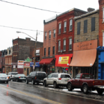 Street view of Beltzhoover in Pittsburgh, featuring storefronts, parked cars, and wet pavement, highlighting the neighborhood's urban environment relevant to local roofing maintenance challenges.
