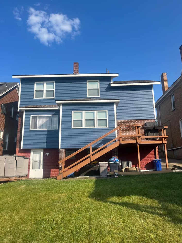 Two-story house with blue siding and brick accents, featuring a wooden staircase leading to a deck, situated on a grassy lawn, highlighting exterior maintenance and curb appeal.