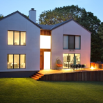 Modern Colonial-style home exterior at dusk, featuring large windows, a wooden deck, and landscaped yard, highlighting architectural beauty and maintenance focus.