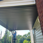 Person on ladder inspecting soffit and fascia under a roof overhang, emphasizing home exterior maintenance and repair.