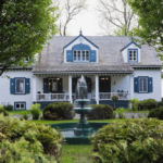 Modern home with blue accents and a fountain, surrounded by greenery, showcasing aesthetic appeal and potential roofing options for Pittsburgh homeowners.