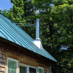 Metal roof with green panels and chimney, showcasing durability and energy efficiency, surrounded by trees, relevant to Pittsburgh roofing solutions.