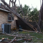 Tree fallen against a house, damaged fence, and debris scattered on the ground, illustrating the aftermath of storm damage and the need for professional roof repair services.