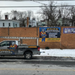 Malick Brothers Exteriors truck parked in front of a brick wall displaying promotional signage for commercial roofing services, including metal roofing installation and maintenance.