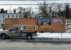 Malick Brothers Exteriors truck parked in front of a commercial building with a snow-covered ground, featuring a prominent sign highlighting roofing services and contact information, relevant to winter roof maintenance in Pittsburgh.