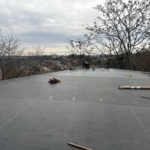 Flat commercial roof installation with tools and materials, overlooking a residential area, emphasizing roofing performance and maintenance considerations.