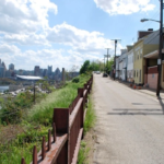 Pittsburgh West End street scene with residential buildings, overgrown grass, and city skyline in the background, reflecting architectural heritage and community resilience.