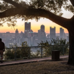 Pittsburgh skyline at sunset with a silhouette of a person standing under a tree, highlighting the city's urban landscape and natural beauty, relevant to durable roofing solutions for homes in Hazelwood.