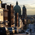 Beechview street scene at sunset with parked cars, a person walking a dog, and prominent church domes in the background, illustrating the neighborhood's charm and potential winter roofing challenges.
