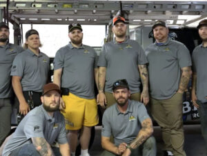 Team of Malick Brothers Exteriors employees in gray shirts, posing in front of their work vehicle, showcasing expertise in roofing and exterior solutions.