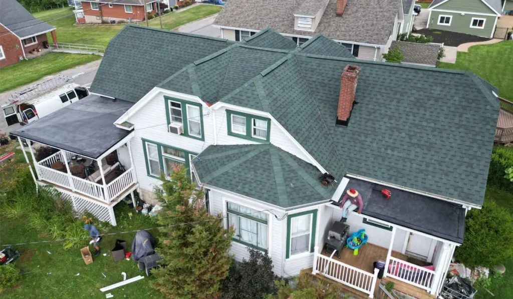 Asphalt shingle roofing installation on a residential home in Pittsburgh, showcasing green shingles, a chimney, and a porch area, with workers and equipment visible.