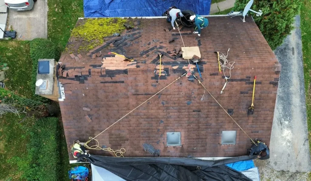 Roofing contractors replacing damaged shingles on a residential roof, showcasing tools, safety ropes, and mossy areas, emphasizing professional roofing services in Pittsburgh.