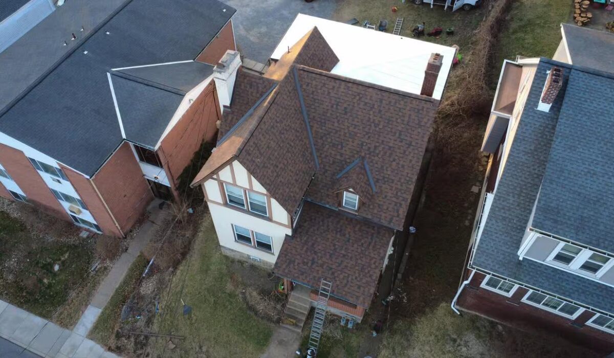 Aerial view of a residential home in Western Pennsylvania showcasing a sloped roof with shingles, adjacent buildings, and a ladder, relevant to gutter guard installation and maintenance discussions.