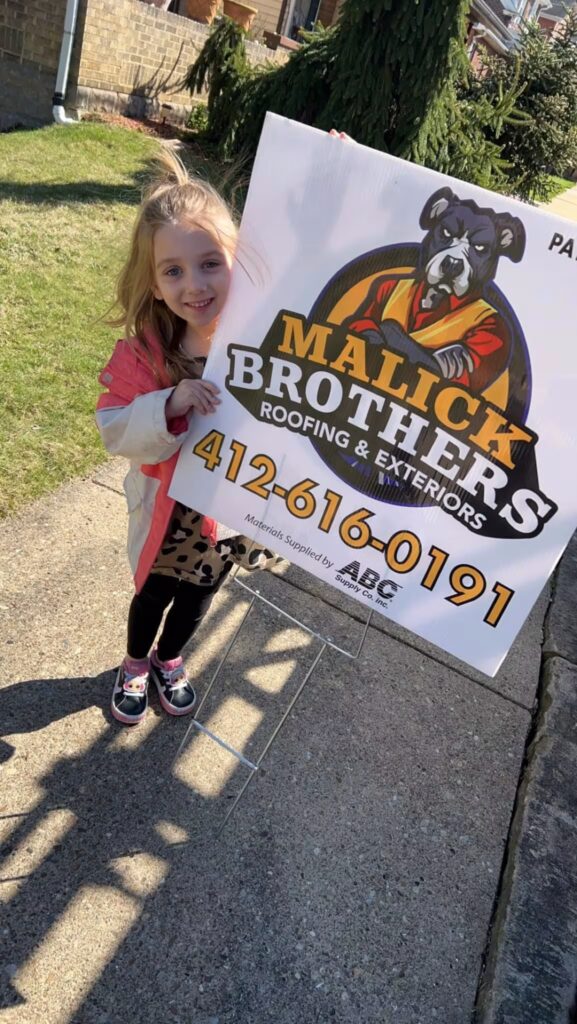Young girl holding a sign for Malick Brothers Roofing & Exteriors, featuring a dog logo and contact number, in a residential neighborhood setting.