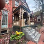 Victorian-style porch with decorative architecture, stairs leading up to the entrance, and yellow flowers in a planter, set in a historic Pittsburgh neighborhood.