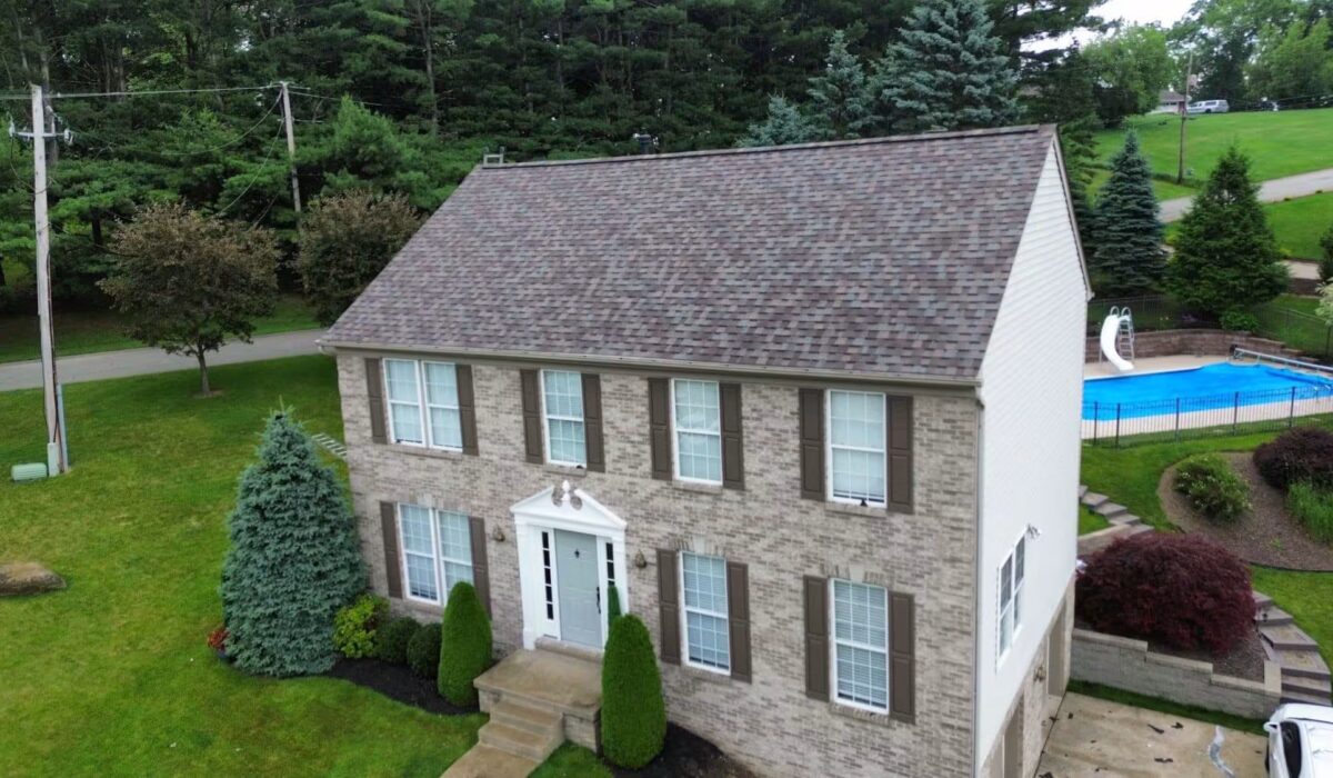 Aerial view of a well-maintained two-story brick home with a new roof and landscaped yard, featuring green shrubs and a swimming pool in the background, illustrating exterior preparation for Memorial Day in Pittsburgh.