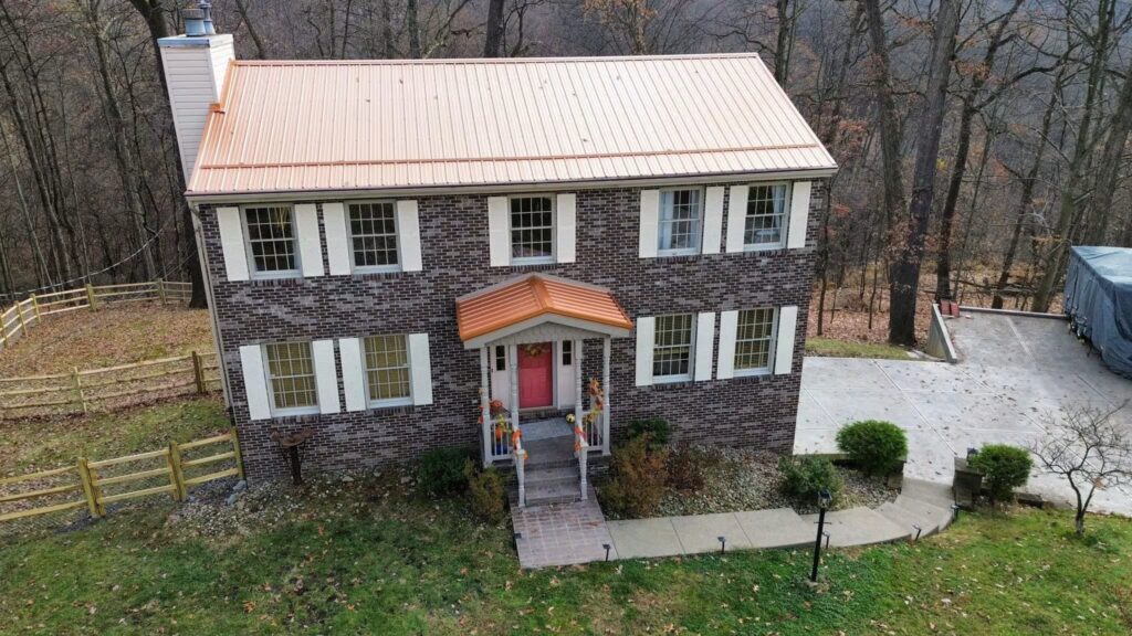 Pittsburgh residential home with brick exterior and orange metal roof, surrounded by trees and a gravel driveway, showcasing preventive roofing solutions.