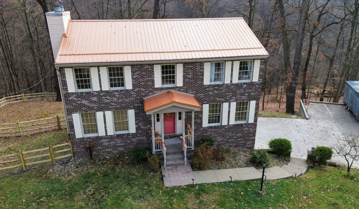 Brick house with a metal roof, featuring a copper-colored front awning and a pink door, surrounded by a landscaped yard and trees, highlighting metal roofing benefits for Pittsburgh homeowners.