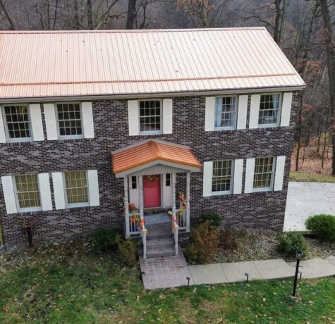 Two-story brick house with a copper-colored metal roof, featuring white window shutters, a pink front door, and a landscaped entrance, showcasing quality exterior craftsmanship relevant to Malick Brothers Exteriors' roofing services.