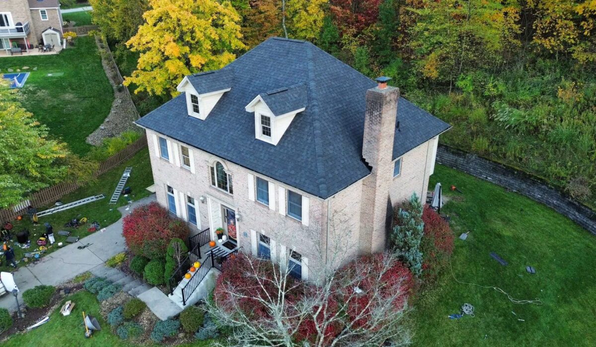 Aerial view of a brick home in Pittsburgh with a newly installed asphalt roof, surrounded by autumn foliage and landscaped yard, illustrating the impact of local weather on roofing durability.