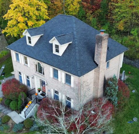 Aerial view of a two-story brick house with a dark shingle roof, surrounded by colorful autumn foliage and landscaped gardens, showcasing quality roofing and exterior features relevant to Malick Brothers Exteriors' services.