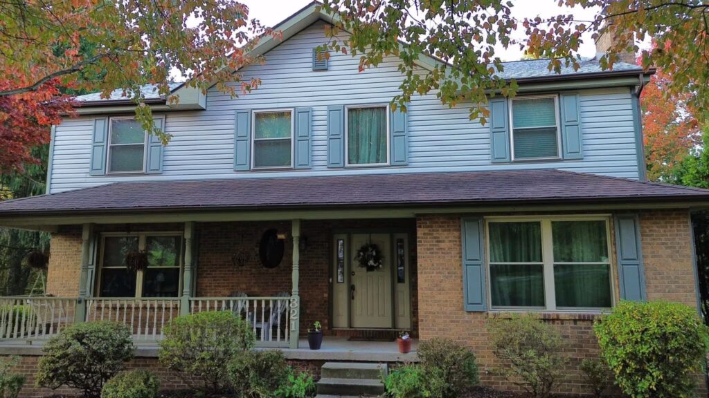 Residential home with a welcoming front porch, featuring a mix of brick and blue siding, surrounded by well-maintained bushes and trees, illustrating durable roofing options for Pittsburgh homeowners.