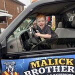 Child smiling and sitting in the driver's seat of a vehicle with "Malick Brothers Exteriors" branding, emphasizing family-oriented service in Pittsburgh roofing.