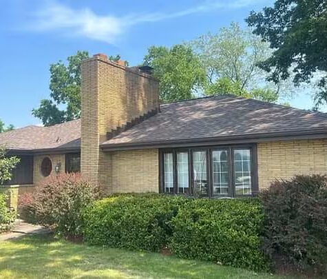 Brick home with a new roof, featuring a prominent chimney and surrounded by well-maintained shrubs and greenery, reflecting quality exterior craftsmanship by Malick Brothers Exteriors.