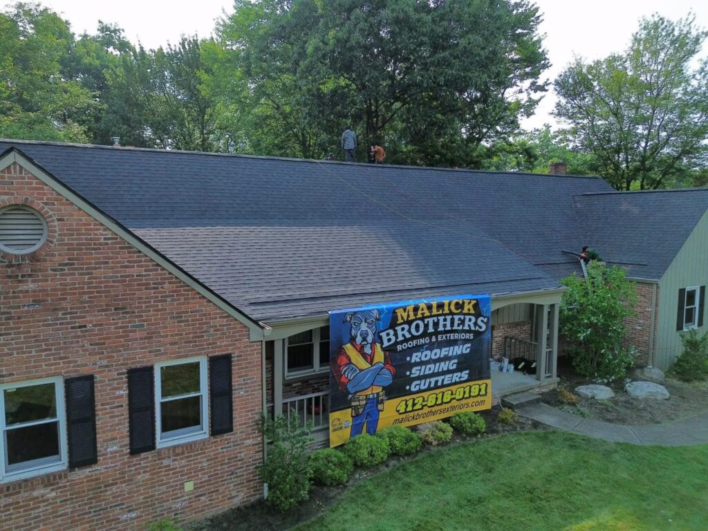 Roofing installation by Malick Brothers Exteriors on a residential home, featuring a new shingle roof, with workers on the roof and a prominent company banner displaying services like roofing, siding, and gutters.