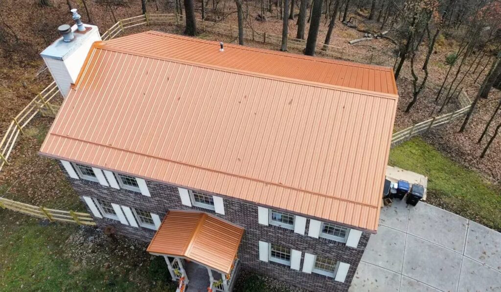 Aerial view of a residential home featuring a new orange metal roof, surrounded by trees and a fenced yard, illustrating roofing options for Pittsburgh homeowners.