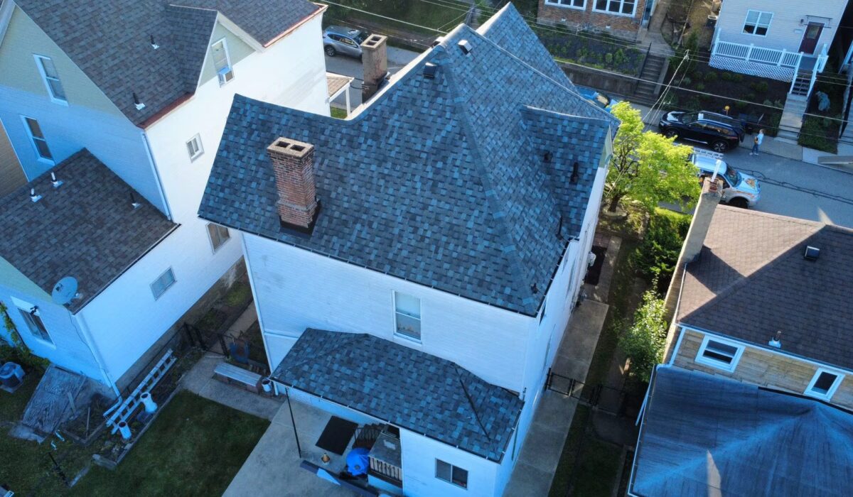 Aerial view of a residential roof in Pittsburgh featuring gray shingles, two chimneys, and adjacent houses, illustrating roofing solutions relevant to homeowners facing repair costs.