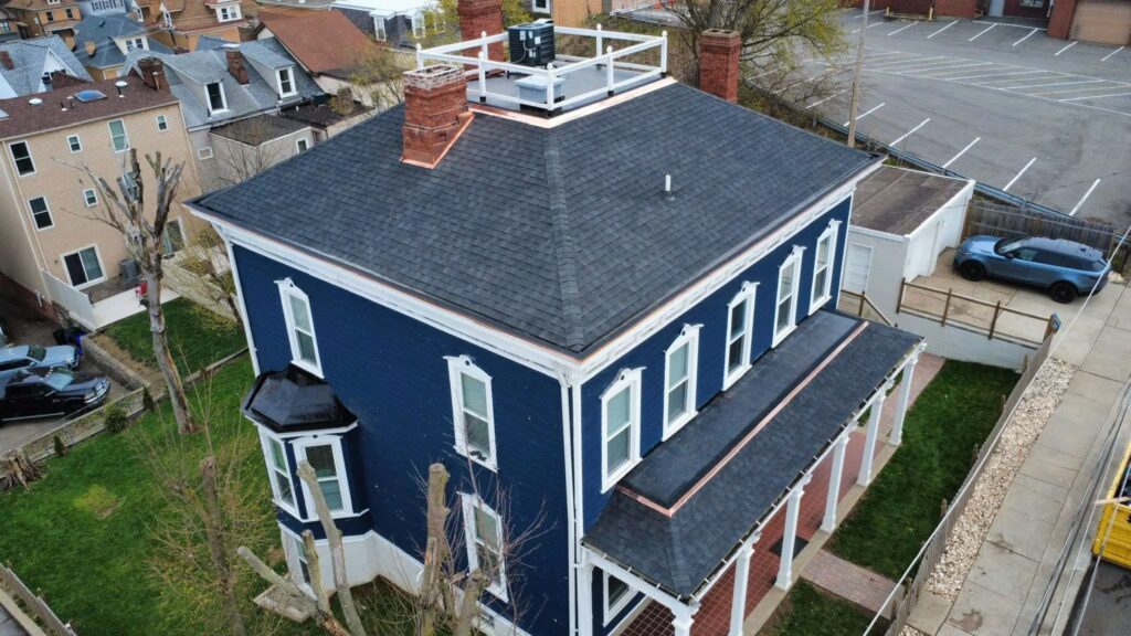 Aerial view of a blue Victorian-style house with a new dark shingle roof, showcasing roofing materials suitable for moisture resistance and climate considerations.