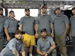 Team of roofing professionals from Malick Brothers Exteriors, wearing branded shirts, posing in front of a work vehicle, showcasing expertise in roofing services.