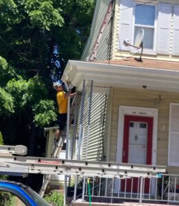 Worker on ladder performing roofing repair on a residential home with green siding and red door, emphasizing the importance of hiring reputable contractors for roofing projects.