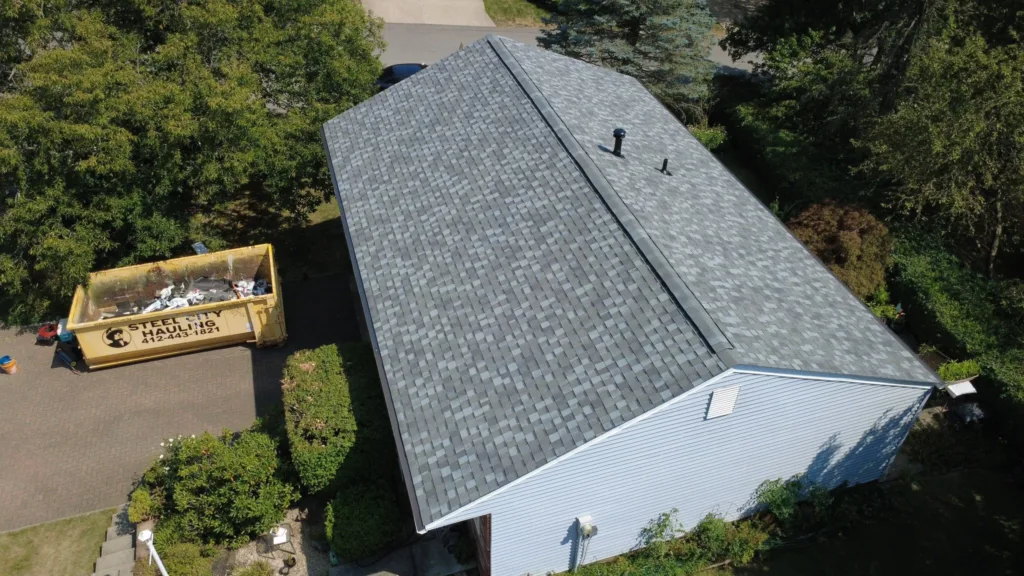 Aerial view of a home in Pittsburgh with a new architectural shingle roof, surrounded by trees and a yellow debris dumpster, emphasizing the importance of proper installation and maintenance for roof longevity.