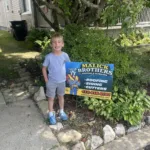 Child standing next to a Malick Brothers Exteriors sign in a residential area, promoting roofing and exterior solutions in Pittsburgh.