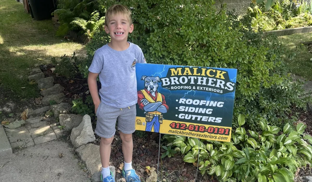 Smiling child standing beside a Malick Brothers Exteriors sign, featuring services for roofing, siding, and gutters, in a residential garden setting.