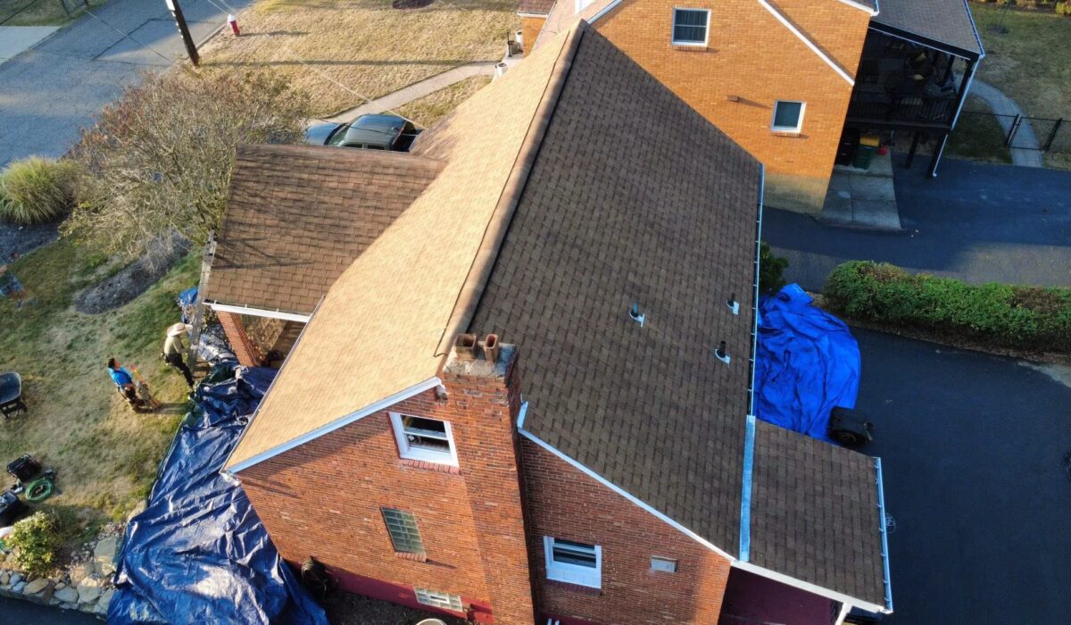 Aerial view of a residential building with a recently installed asphalt shingle roof, workers on the ground near blue tarps, and surrounding greenery, relevant to commercial roofing warranty discussions in Pittsburgh.
