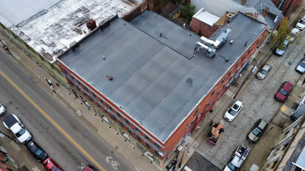 Aerial view of a commercial building with a flat roof, featuring gray roofing material, adjacent streets with parked cars, and surrounding brick structures, illustrating the context of commercial roofing maintenance and investment strategies.