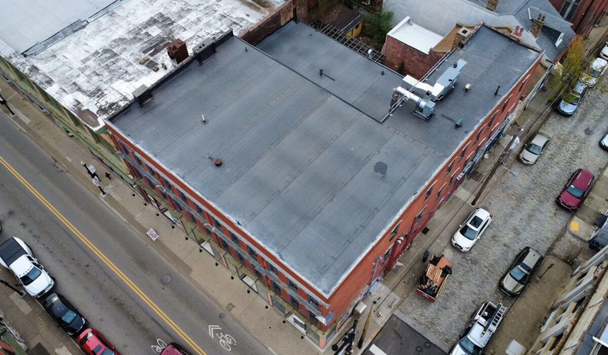 Aerial view of a commercial flat roof with dark membrane, surrounded by urban buildings and parked cars, illustrating roofing material options for commercial structures.