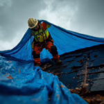 Roofer installing a blue tarp over a damaged shingle roof before stormy weather